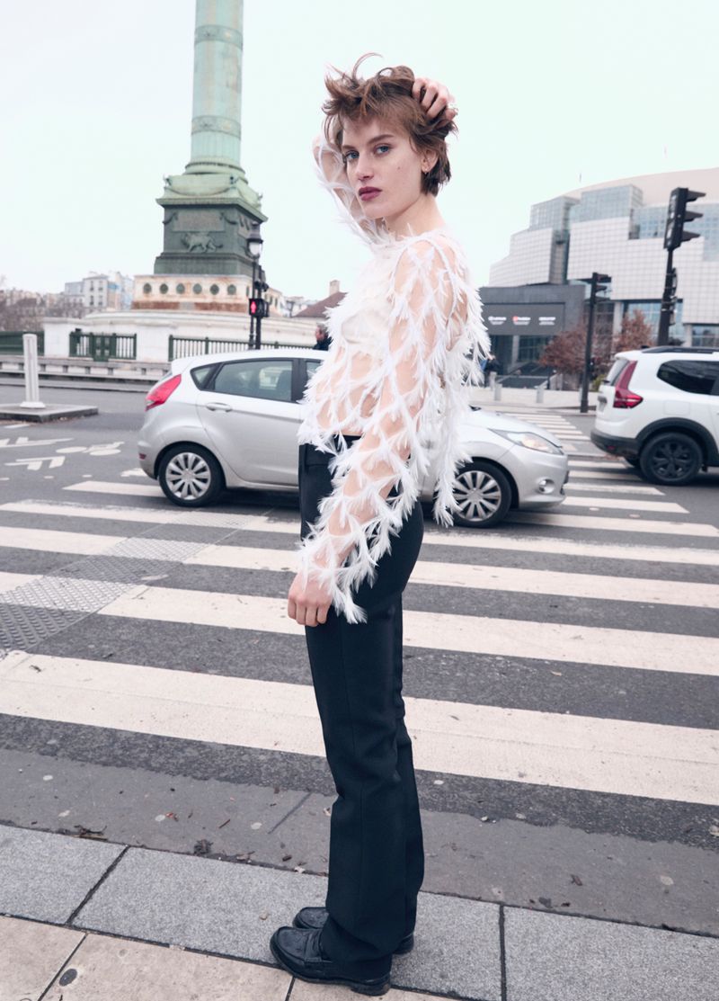 Fashion model poses in a white, feathered top and black pants on a city crosswalk in Paris, with cars and a monument in background.