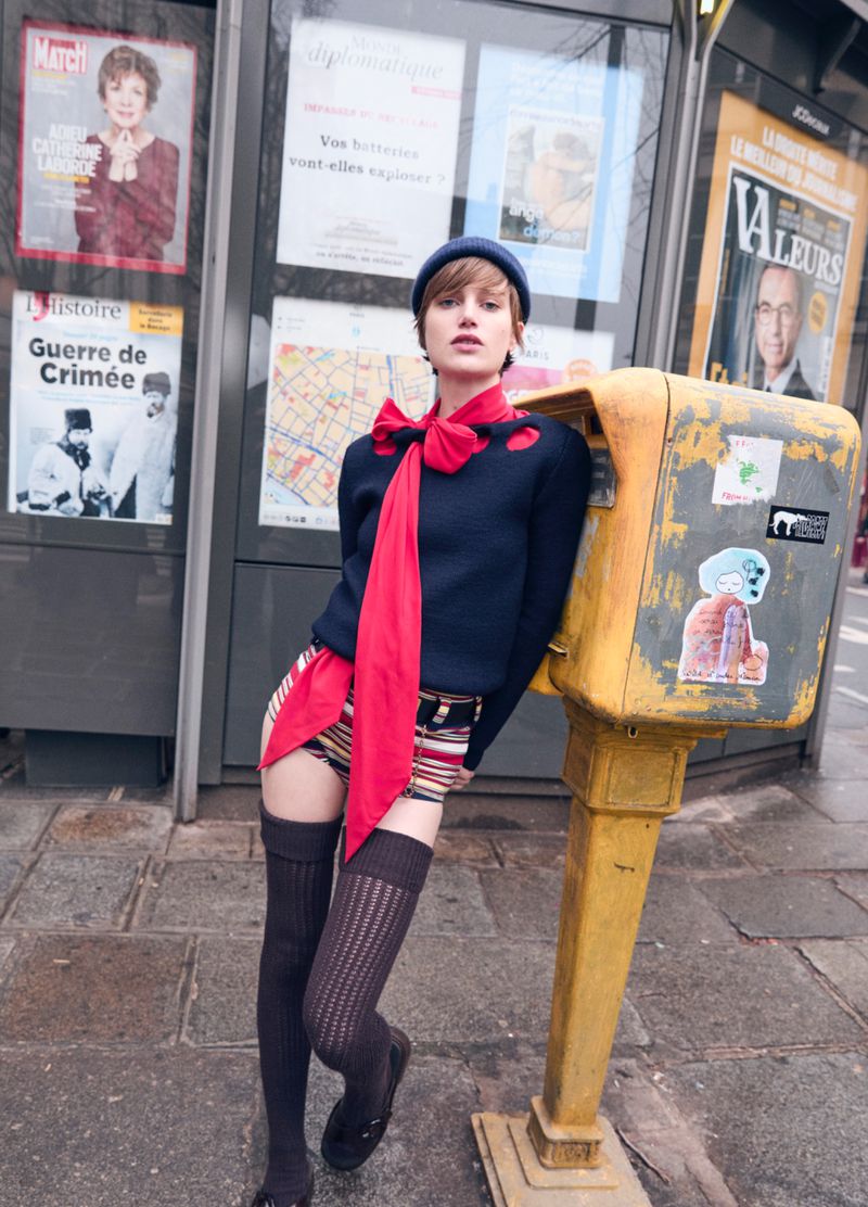 A young adult female model poses in an urban setting wearing a blue sweater, striped shorts, knee-high socks, and a matching blue beret.