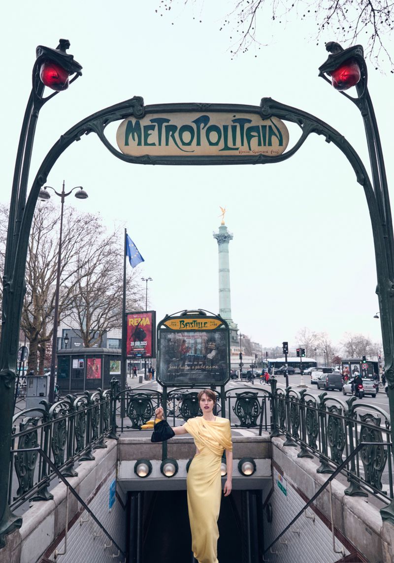 Woman poses at the Paris Métropolitain entrance in a draped yellow dress and black purse, with a view of the city street beyond.