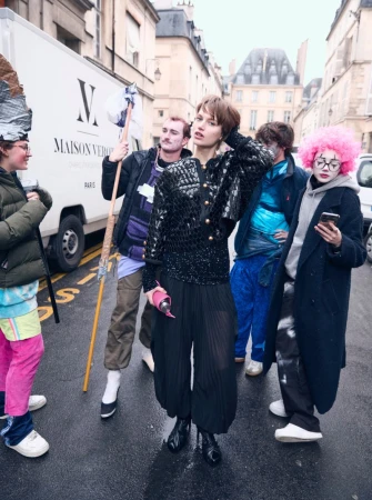 Fashion photo shoot on a city street featuring a model wearing a sequined top and balloon pants, surrounded by a group of people.