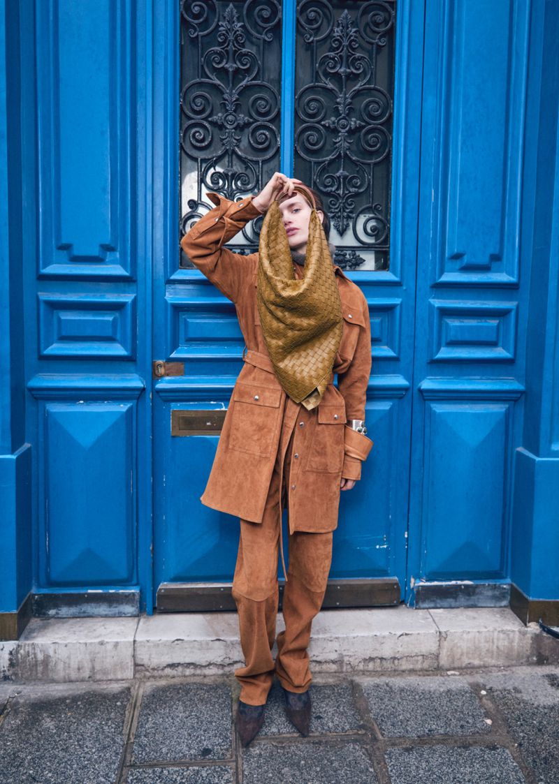 Full-length shot of a slender person in a tan outfit and patterned scarf, posing in front of a dark blue door with ironwork.
