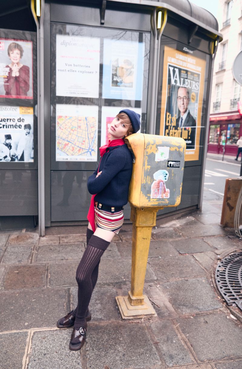 Young adult in beret, school uniform, and knee-highs leans against yellow post box near bus stop in Paris with posters.