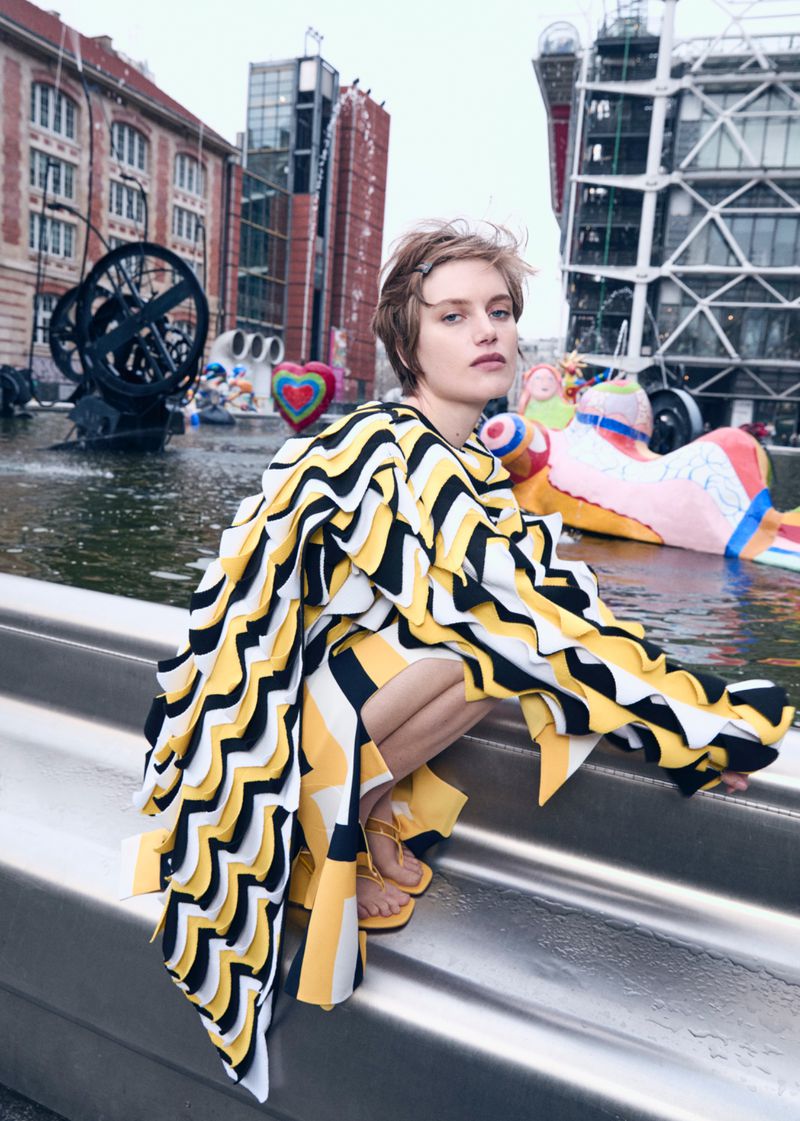 A woman with short blonde hair wearing a black and yellow striped coat crouching on stairs outdoors in an urban setting with architecture.