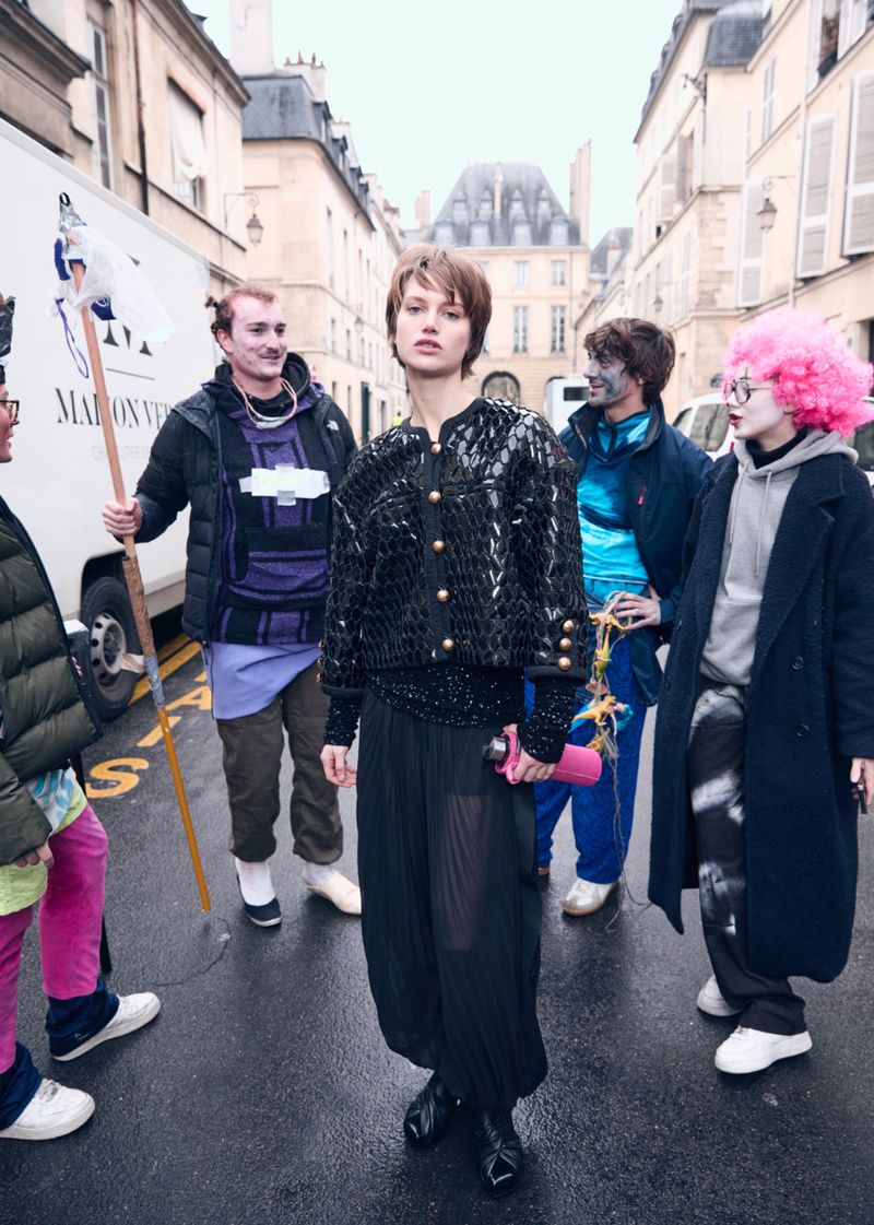 Fashion shot on a street with a woman as the main subject, wearing a sequin jacket and sheer black pants, surrounded by a group.