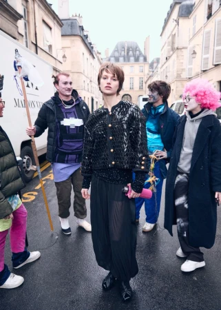 Fashion shot on a street with a woman as the main subject, wearing a sequin jacket and sheer black pants, surrounded by a group.