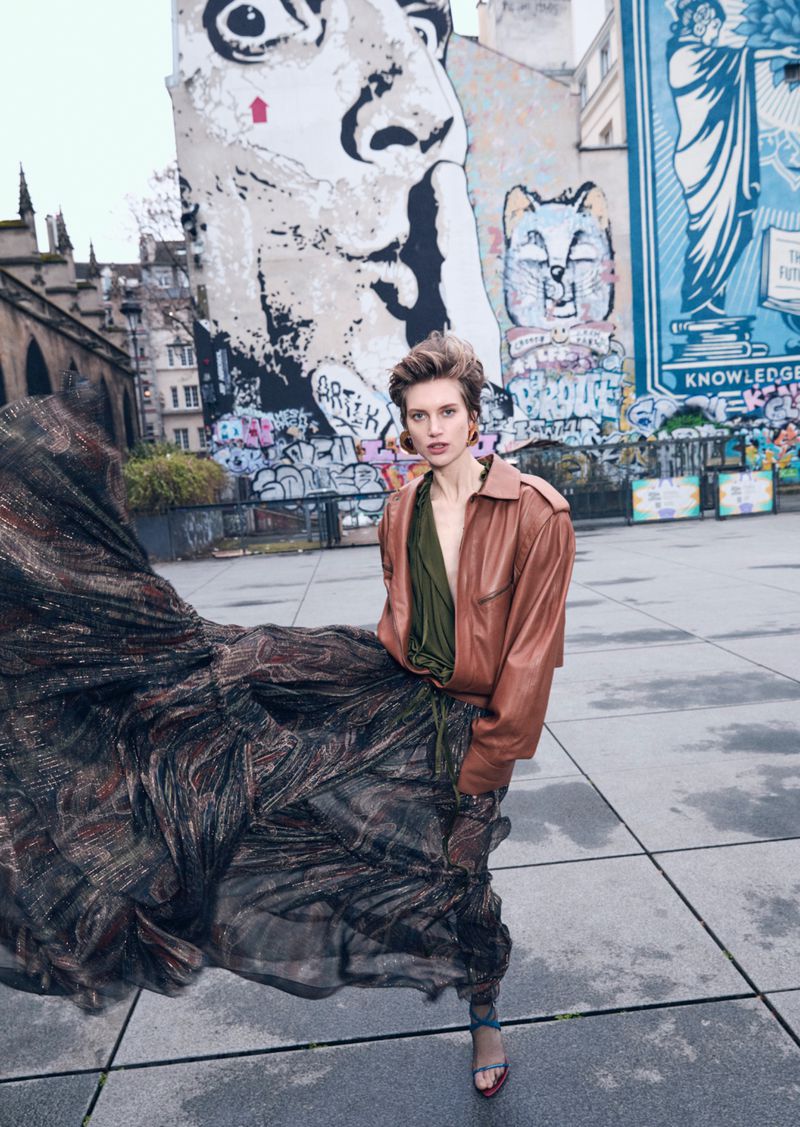 Fashion shot of a woman in a tan jacket and flowing skirt, standing on a gray plaza with a large mural and buildings in the background.
