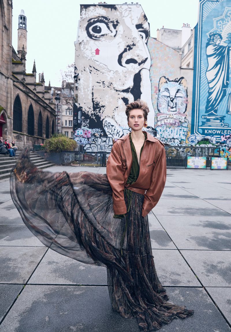 A woman in a brown leather jacket and long skirt poses on an outdoor plaza with street art on buildings in the background.
