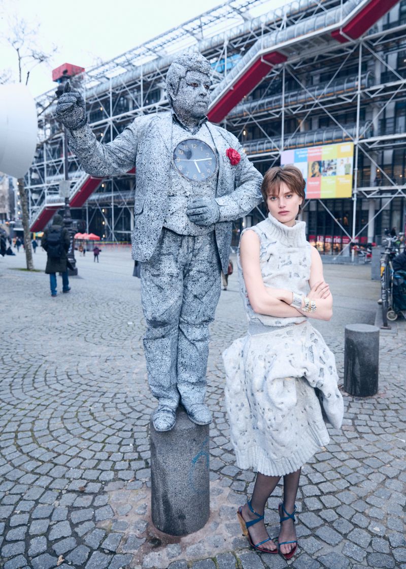 Fashionable woman poses next to a human statue in a grey dress and heels in an urban square with modern building in the background.