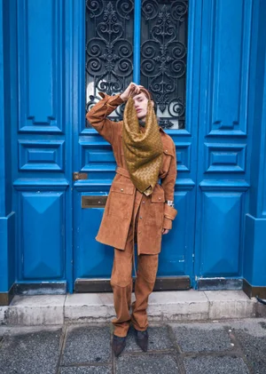 Model wearing a brown suede jacket and matching trousers posing in front of a large vibrant blue wooden door with intricate black ironwork.
