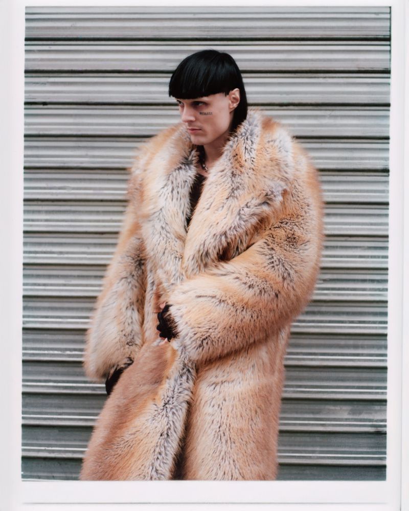Person posing outdoors against a grey roll-up door wearing a large, light brown fur coat with a black bowl cut hairstyle.
