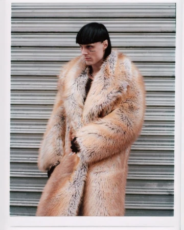 Person posing outdoors against a grey roll-up door wearing a large, light brown fur coat with a black bowl cut hairstyle.