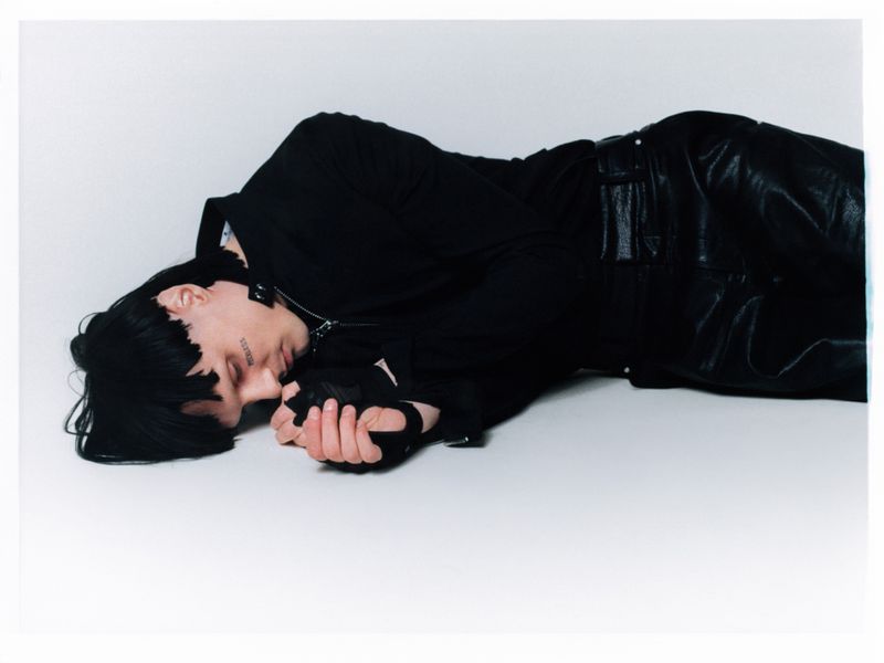 Lying down studio shot of a person with black hair, wearing all black, on a white background. Silver necklace. Studio lighting.