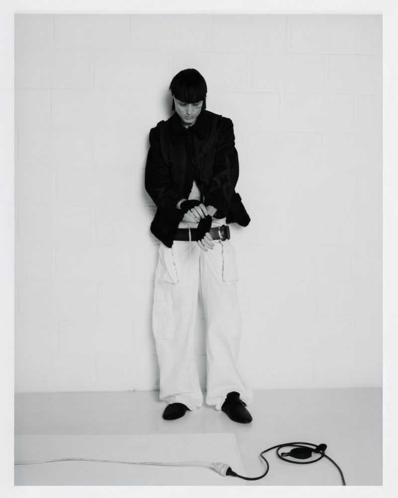 Monochromatic portrait of a person in a black jacket and white pants standing in front of a white wall, adjusting their sleeve cuffs.