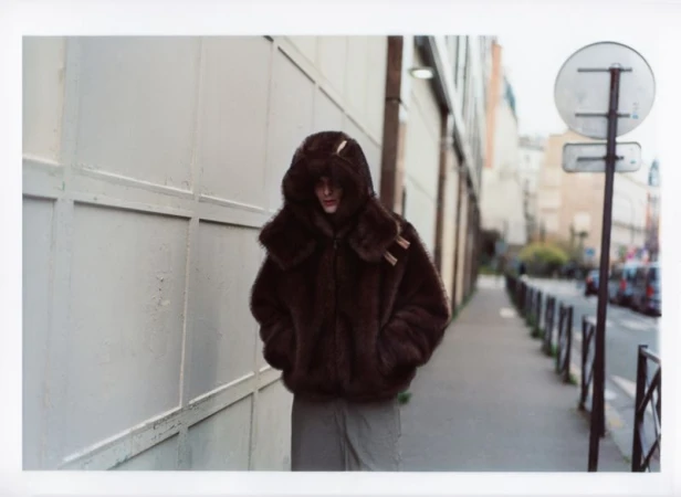 An adult in a brown faux fur hooded coat stands on a sidewalk next to a light gray wall, on an urban street with cars in the background.