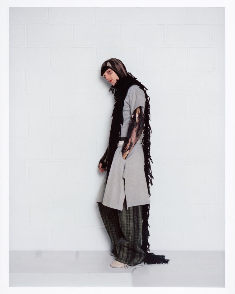 Portrait of a man wearing a grey tunic, patterned trousers, and a long black fringed scarf against a white wall.