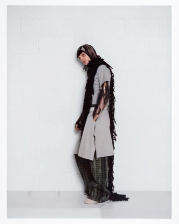Portrait of a man wearing a grey tunic, patterned trousers, and a long black fringed scarf against a white wall.