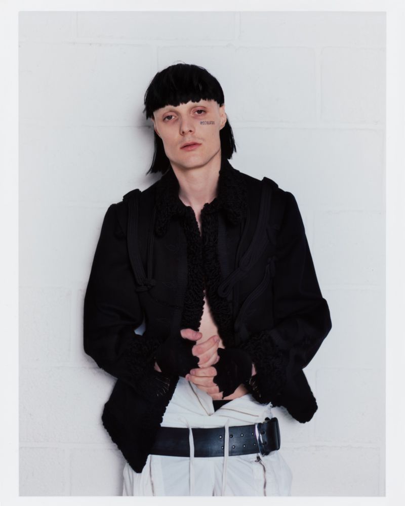 Portrait of an adult male with a visible tattoo under his eye, wearing a black jacket and white pants, posing against a white wall.