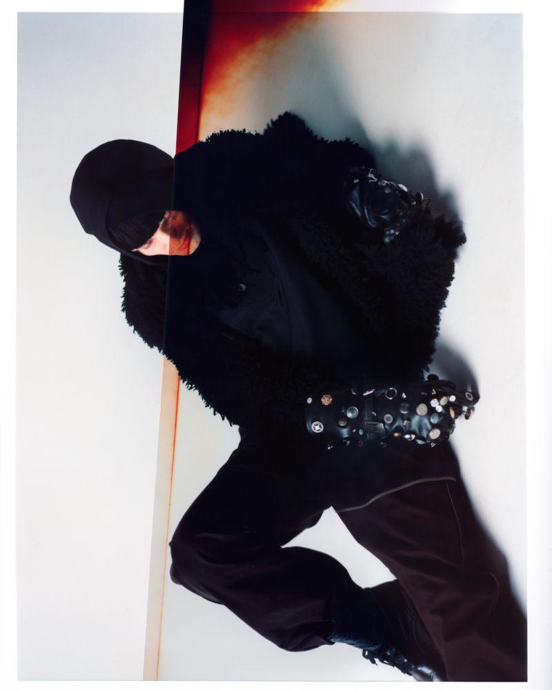 Fashion shot of a person wearing a black furry jacket, black hat, and brown pants, posing indoors against a light-colored backdrop.
