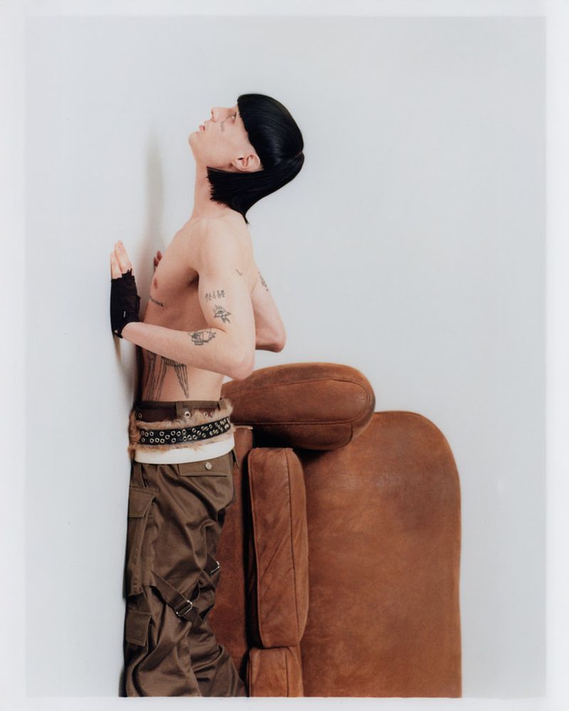Portrait of a shirtless person with a bowl cut and tattoos leaning against a white wall next to a brown leather chair indoors.
