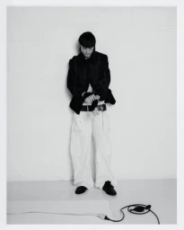 Model standing against a white wall wearing a dark jacket and light trousers for Office Magazine photographed by Letizia Guel.