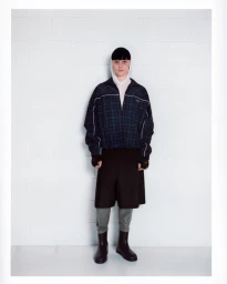 Full-length portrait of a person wearing a dark plaid zip jacket, white hoodie, dark shorts, and boots against a white brick wall.