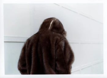 A person wearing a textured brown faux fur hooded jacket standing against a white paneled wall.