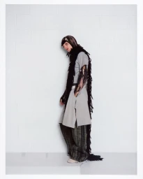 Portrait of a man wearing a grey tunic, patterned trousers, and a long black fringed scarf against a white wall.