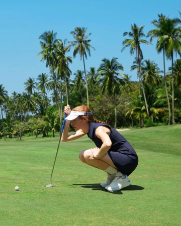 A woman wearing golf attire is crouched down on a golf course with a putter, lining up a putt on a bright sunny day.