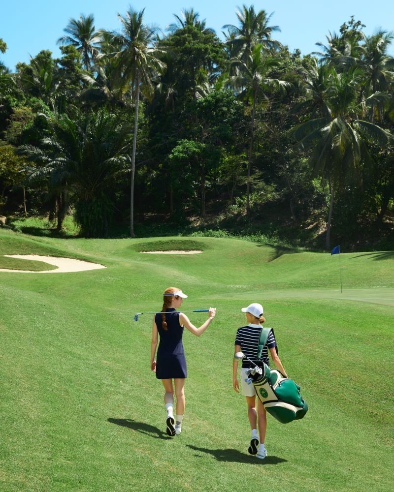 Two adult females walk on a golf course with trees in the background; one holds a golf club and the other carries a golf bag.
