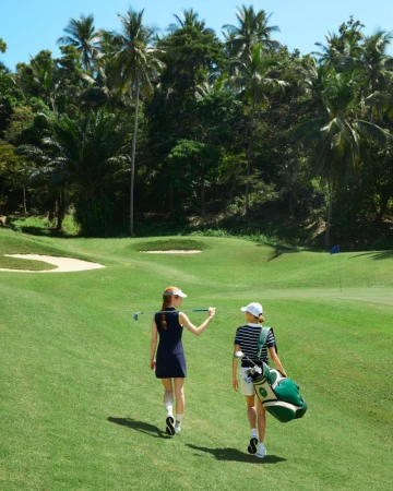 Two adult females walk on a golf course with trees in the background; one holds a golf club and the other carries a golf bag.