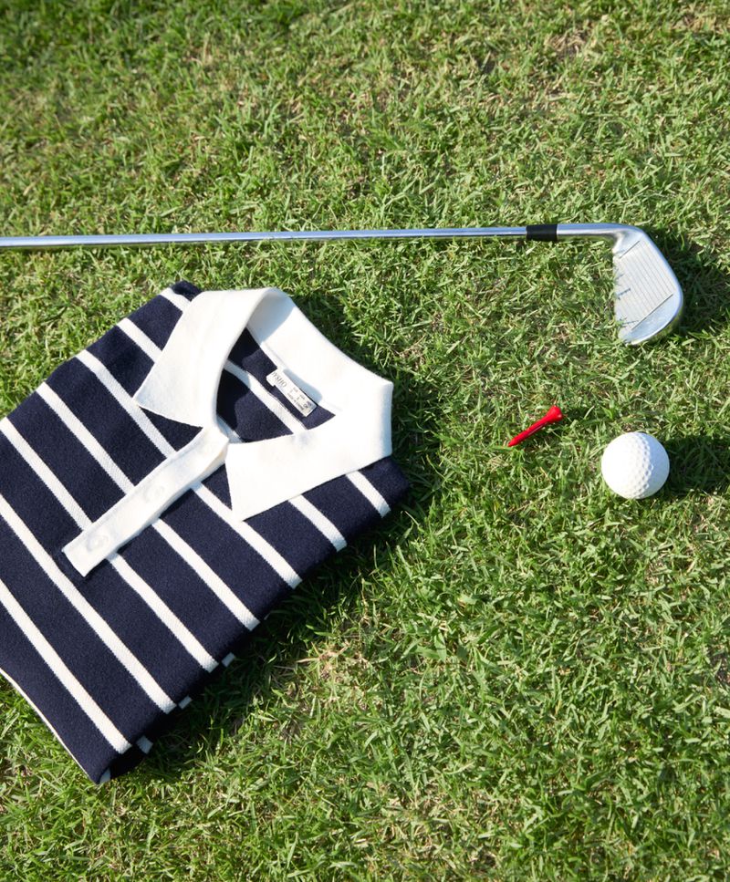 Golf polo shirt and golf equipment on grass. Striped golf shirt with collar, tee, golf ball, and golf club on green grass.