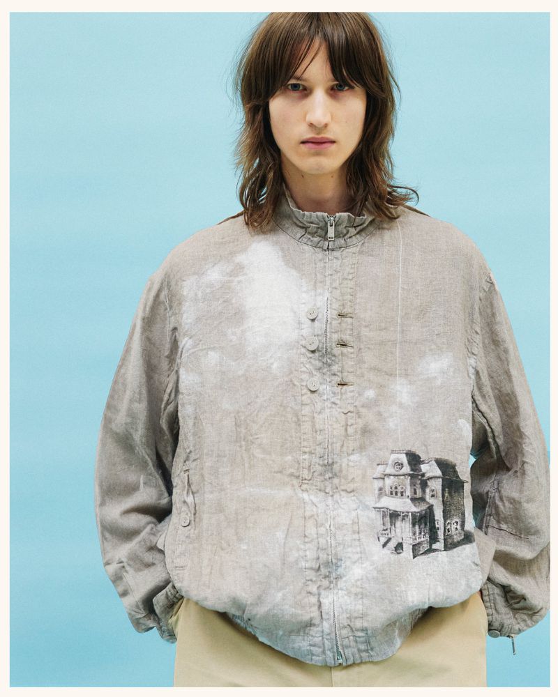Male model wearing a grey jacket with a house print against a blue background. Fashion shot showcasing unique jacket design and neutral colors.