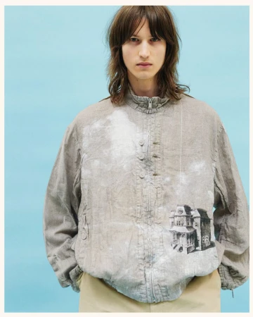 Male model wearing a grey jacket with a house print against a blue background. Fashion shot showcasing unique jacket design and neutral colors.