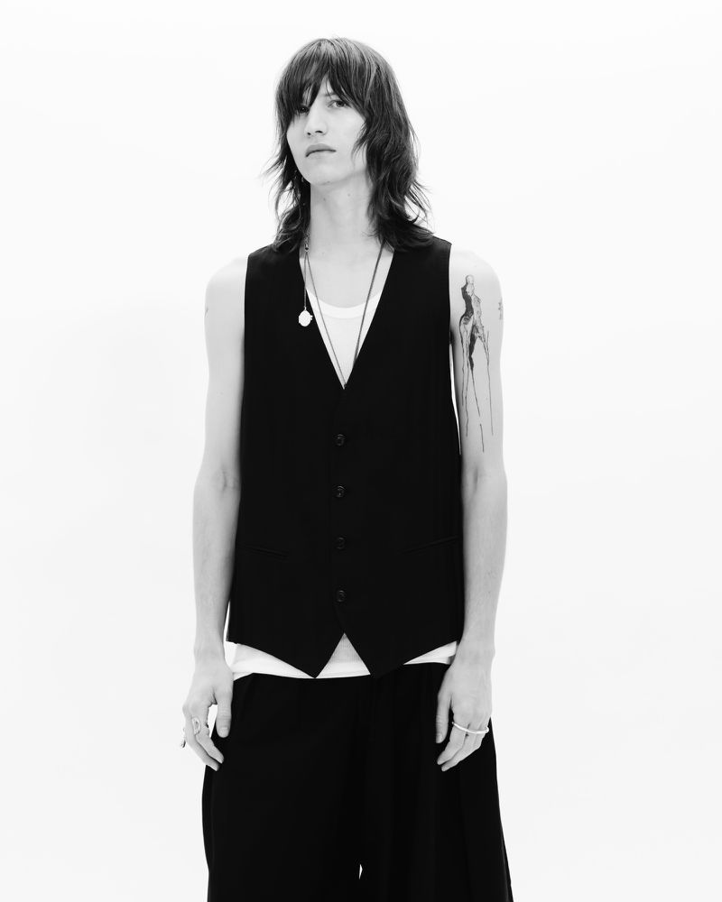 Black and white portrait of a slender person with long hair wearing a black vest, set against a bright white backdrop in a studio setting.