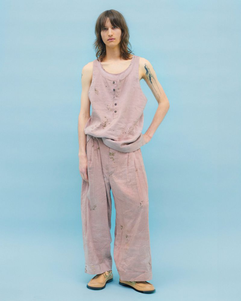 Full-body portrait of a young adult wearing a rose-colored linen tank top and matching wide-leg pants against a light blue background.