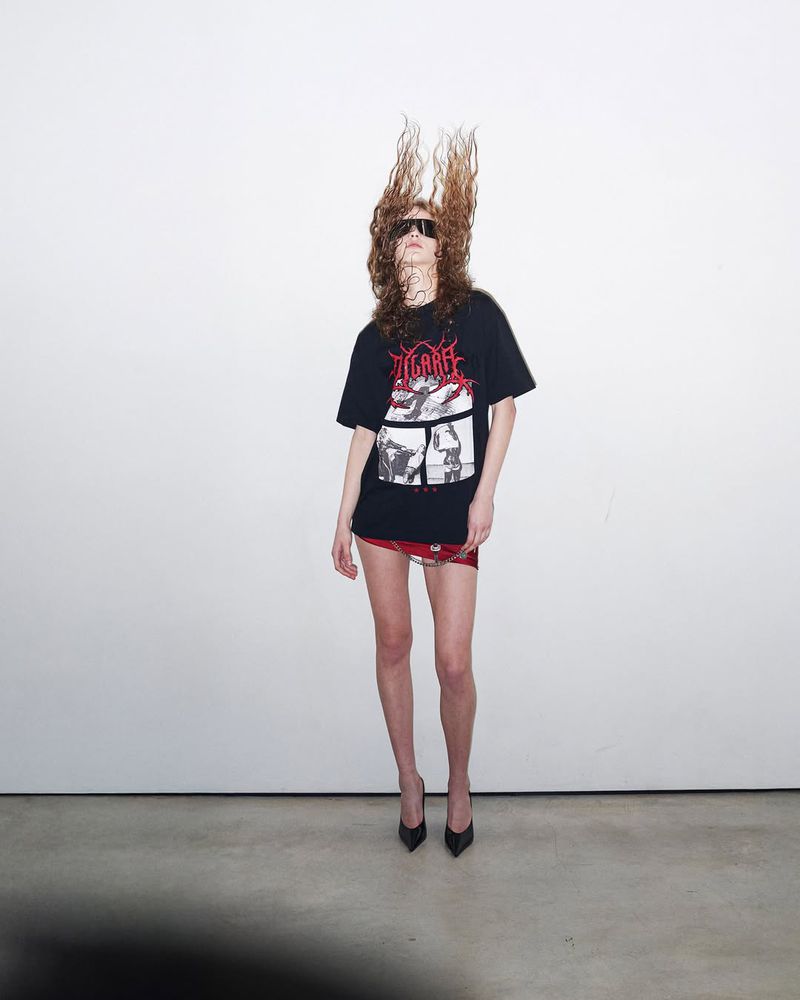 Woman with wavy brown hair wears a graphic tee, red miniskirt, and heels, standing against a white wall in a studio.