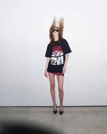 Woman with wavy brown hair wears a graphic tee, red miniskirt, and heels, standing against a white wall in a studio.