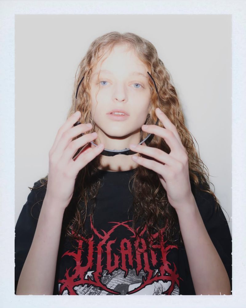 Young adult woman with wavy hair poses indoors wearing a graphic tee shirt and choker, looking directly at the camera.