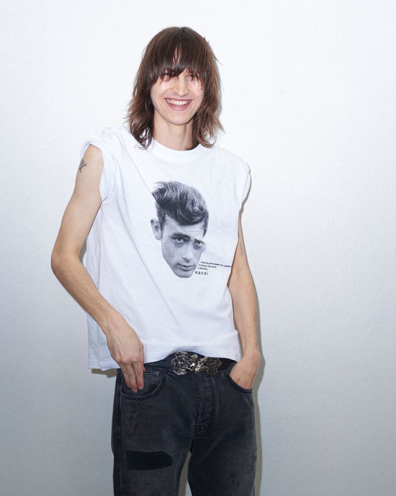 Adult male poses in a white sleeveless t-shirt with a black and white print of a male face, wearing grey jeans with a large patterned belt.