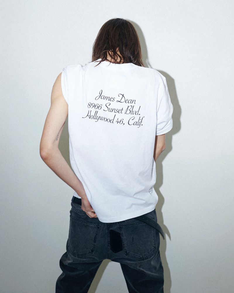Model with long brown hair seen from behind wearing a white t-shirt and dark jeans against a white wall.