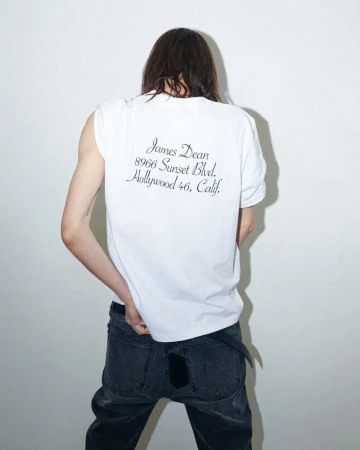 Model with long brown hair seen from behind wearing a white t-shirt and dark jeans against a white wall.
