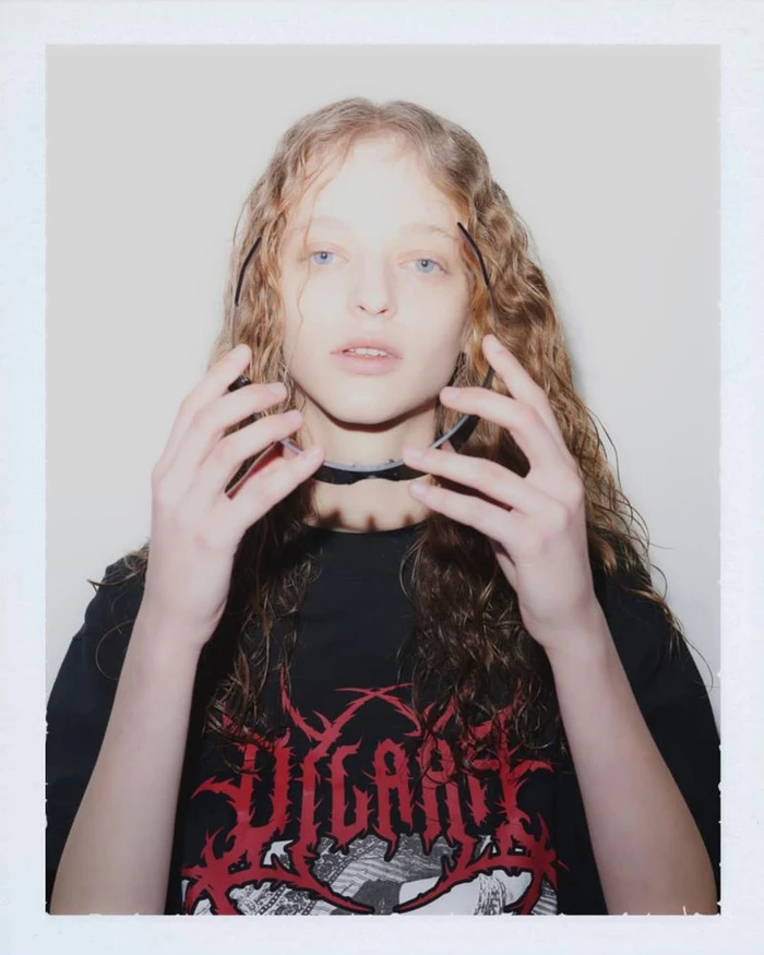 Portrait of a young model with curly hair holding black sunglasses against a white background.