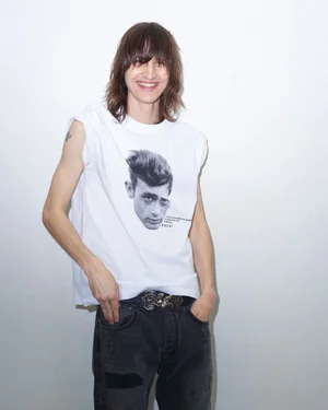 A smiling male model with long brown hair wearing a white graphic t-shirt and dark jeans against a plain white background.