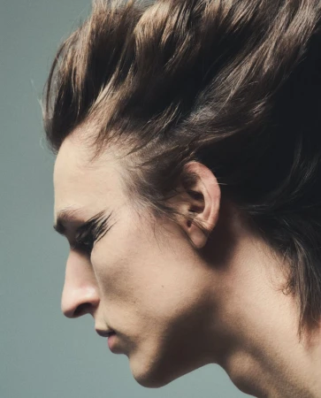 Close-up profile of a model with dramatic black eye makeup and wind-blown brown hair against a neutral gray background
