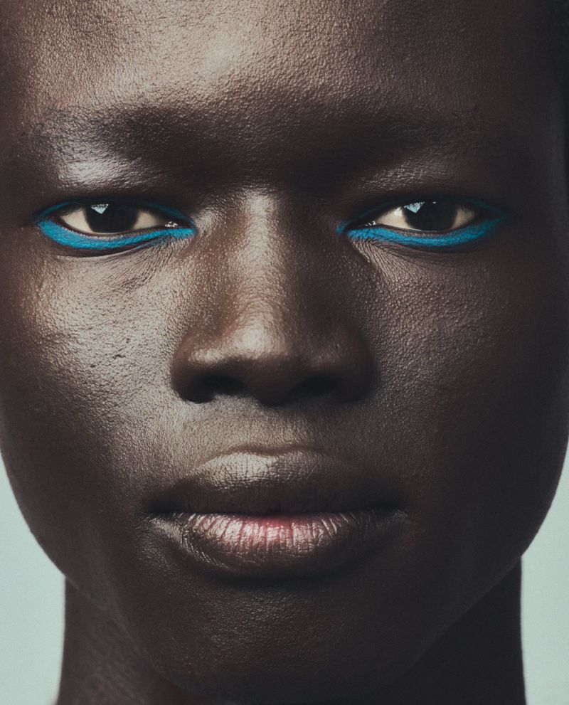 Close-up portrait of an adult woman with dark skin, blue eyeliner, and glossy lips against a light blue background in a beauty context.