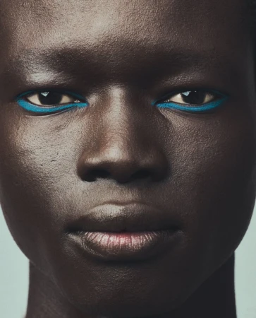 Close-up portrait of an adult woman with dark skin, blue eyeliner, and glossy lips against a light blue background in a beauty context.