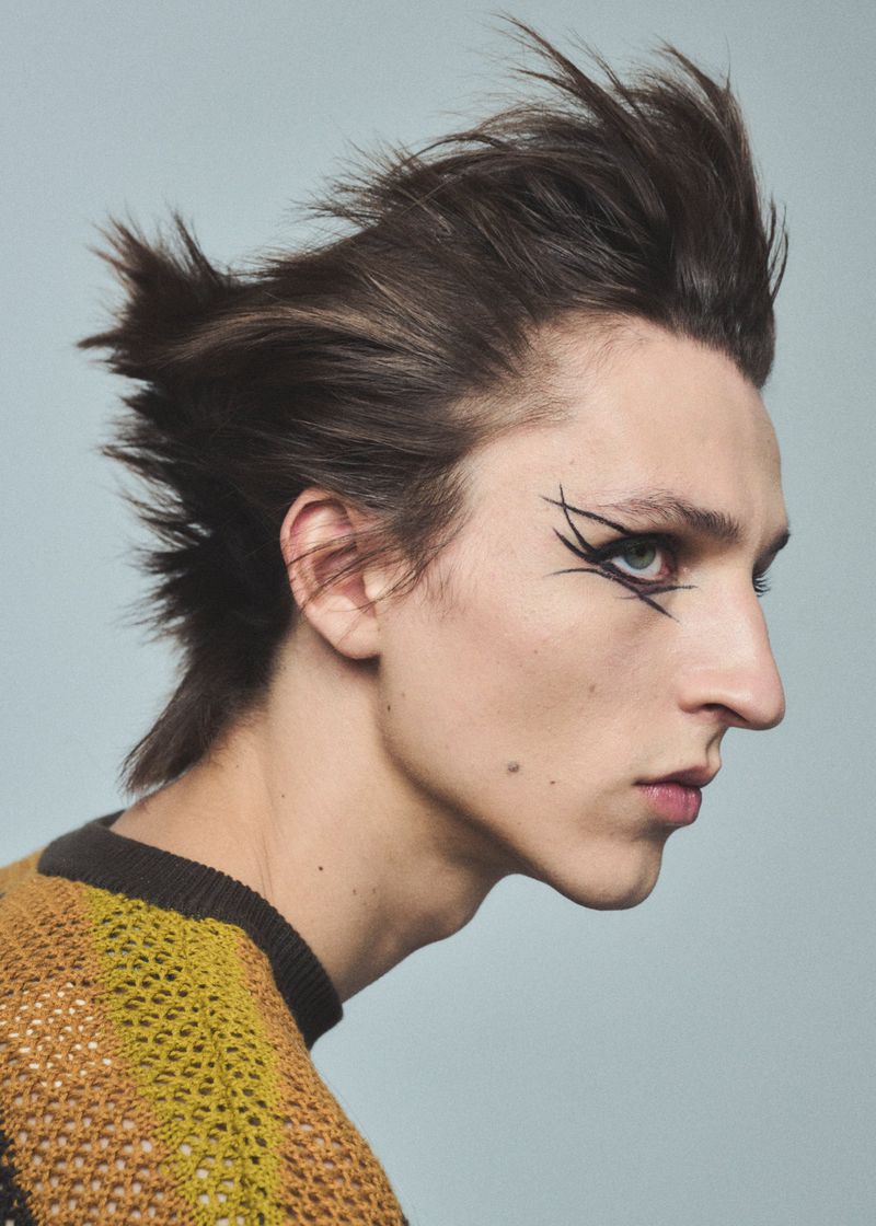 Portrait of a young adult with brown hair styled up and back, wearing eye makeup, in a yellow-brown knit top, against a light blue background.