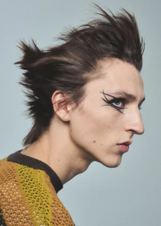Portrait of a young adult with brown hair styled up and back, wearing eye makeup, in a yellow-brown knit top, against a light blue background.