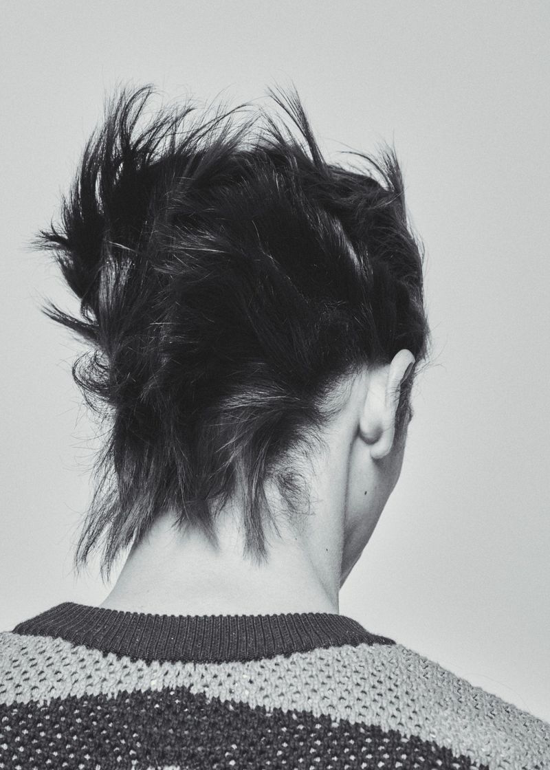 Rear view of a person with short textured dark hair and a patterned knit sweater against a plain neutral background.