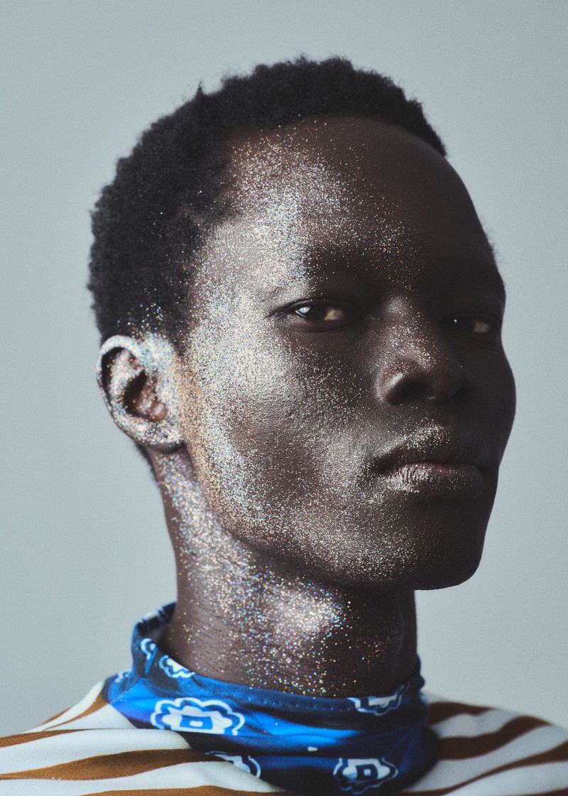 Close-up studio portrait of a dark-skinned adult male with glitter on his face and neck, wearing a striped shirt and blue bandana.